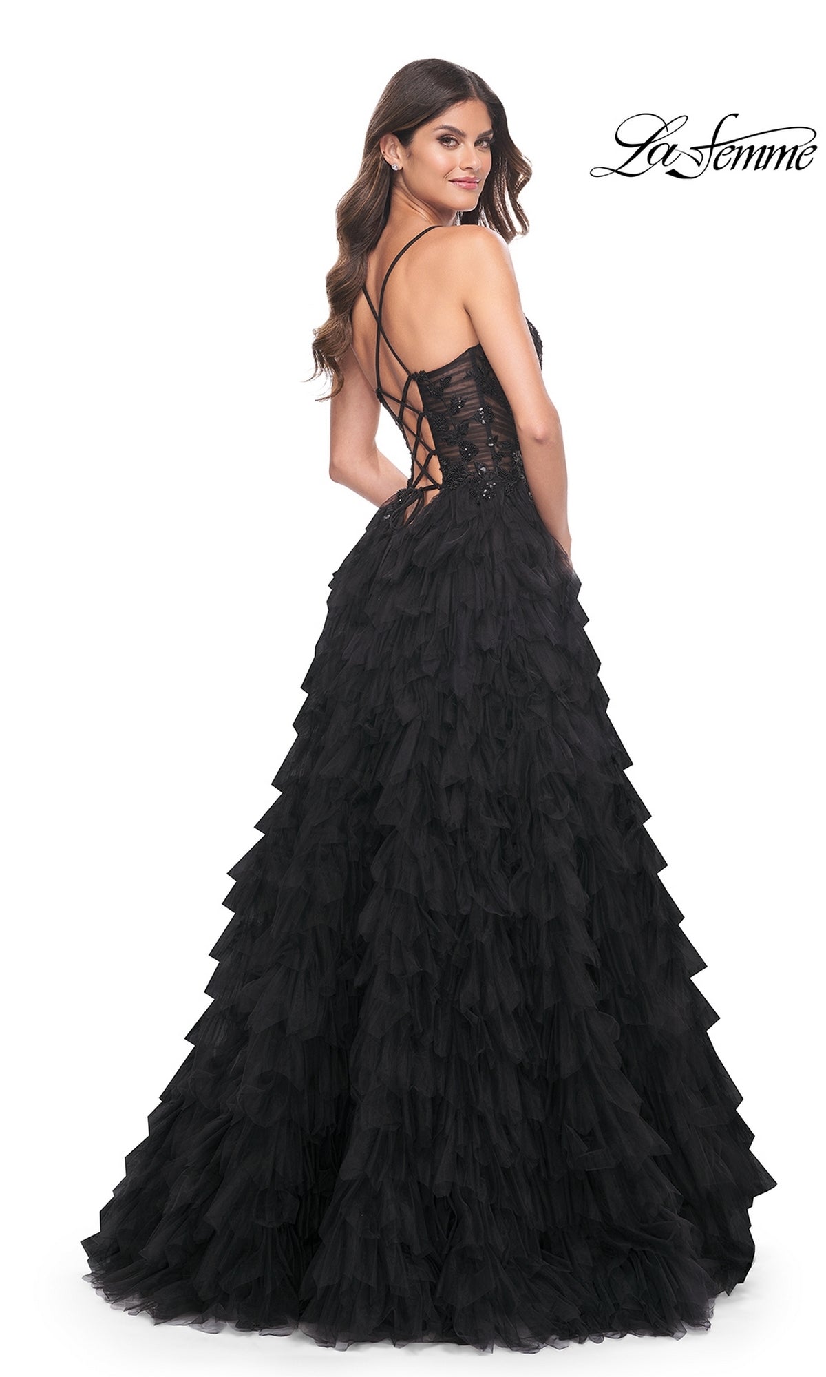 A woman models the La Femme Long Black Ruffled Prom Ball Gown 32108, featuring a lace-up open back, as she stands looking over her shoulder. The La Femme logo appears in the upper right corner.
